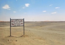A sparse desert landscape with a directional signpost indicating camping facilities 1500 meters ahead. The sign reads 'Camping Secret de Sahara' and mentions a caf&eacute; and restaurant. The horizon features distant sandy dunes under a clear blue sky with a few scattered clouds.