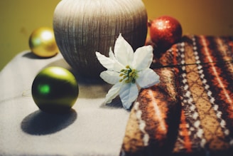 A decorative scene featuring a white flower resting on a patterned brown and orange textile, accompanied by a light wood-colored vase. Surrounding the arrangement are Christmas ornaments in gold and green, adding a festive touch.