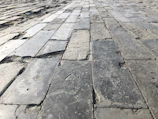 A close-up perspective of an uneven stone pavement, with rectangular stones laid out in a grid pattern. The surface has a mix of smoother and rougher textures with visible wear and aging, signifying use over time.