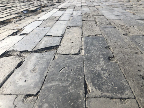 A close-up perspective of an uneven stone pavement, with rectangular stones laid out in a grid pattern. The surface has a mix of smoother and rougher textures with visible wear and aging, signifying use over time.