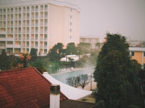 A multi-story beige hotel building with arched windows sits behind lush greenery and palm trees. In the foreground, a red-tiled roof is visible, along with a white structure resembling a canopy. A pool, partially obscured by mist, is surrounded by white loungers and flanked by trees.