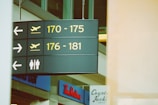 A directional sign in an airport, indicating gate numbers 170 to 175 and 176 to 181, with arrows and airplane symbols. The sign also shows icons for male and female restrooms. Below the sign, various storefronts such as Tim Hortons and Coyote Jack's Grill are partially visible.