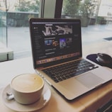 A laptop displaying a music streaming service with a playlist open on the screen. Next to the laptop, there is a white cup filled with frothy coffee on a saucer with a teaspoon. The setup is on a table near a large window, providing a view of the outside with some greenery.