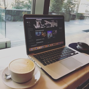 A laptop displaying a music streaming service with a playlist open on the screen. Next to the laptop, there is a white cup filled with frothy coffee on a saucer with a teaspoon. The setup is on a table near a large window, providing a view of the outside with some greenery.