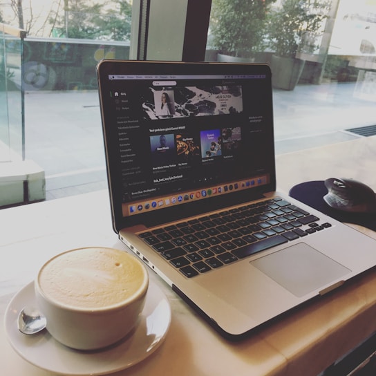 A laptop displaying a music streaming service with a playlist open on the screen. Next to the laptop, there is a white cup filled with frothy coffee on a saucer with a teaspoon. The setup is on a table near a large window, providing a view of the outside with some greenery.