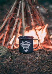 A black enamel mug with the words 'The Adventure Begins' is placed on the ground, surrounded by soil and pine needles. In the background, a campfire is burning brightly, with a teepee-like arrangement of sticks supporting the flames.