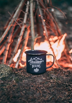 A black enamel mug with the words 'The Adventure Begins' is placed on the ground, surrounded by soil and pine needles. In the background, a campfire is burning brightly, with a teepee-like arrangement of sticks supporting the flames.
