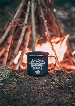 A black enamel mug with the words 'The Adventure Begins' is placed on the ground, surrounded by soil and pine needles. In the background, a campfire is burning brightly, with a teepee-like arrangement of sticks supporting the flames.