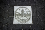 A stone plaque set into a dark cobblestone path features intricate engravings. The central image displays a circular emblem with a cow, corn, and forest trees. Below, the words 'Vermont Freedom Unity' are prominently carved.