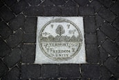 A stone plaque set into a dark cobblestone path features intricate engravings. The central image displays a circular emblem with a cow, corn, and forest trees. Below, the words 'Vermont Freedom Unity' are prominently carved.
