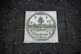 A stone plaque set into a dark cobblestone path features intricate engravings. The central image displays a circular emblem with a cow, corn, and forest trees. Below, the words 'Vermont Freedom Unity' are prominently carved.