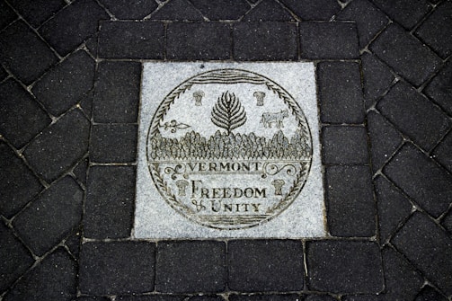 A stone plaque set into a dark cobblestone path features intricate engravings. The central image displays a circular emblem with a cow, corn, and forest trees. Below, the words 'Vermont Freedom Unity' are prominently carved.