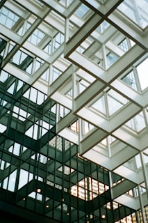 An architectural structure features intersecting glass panels and geometric metal framework. The design creates a complex pattern of reflective surfaces and angular lines. Natural light filters through the windows, casting intricate shadows and reflections.