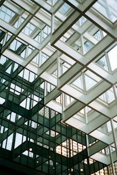 An architectural structure features intersecting glass panels and geometric metal framework. The design creates a complex pattern of reflective surfaces and angular lines. Natural light filters through the windows, casting intricate shadows and reflections.
