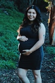 A pregnant woman wearing a dark dress smiles while standing on a pathway surrounded by lush greenery. Her hands gently rest on her belly, and she has long, wavy hair illuminated by sunlight.