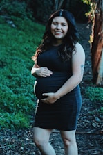 A pregnant woman wearing a dark dress smiles while standing on a pathway surrounded by lush greenery. Her hands gently rest on her belly, and she has long, wavy hair illuminated by sunlight.