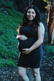 A pregnant woman wearing a dark dress smiles while standing on a pathway surrounded by lush greenery. Her hands gently rest on her belly, and she has long, wavy hair illuminated by sunlight.