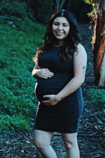 A pregnant woman wearing a dark dress smiles while standing on a pathway surrounded by lush greenery. Her hands gently rest on her belly, and she has long, wavy hair illuminated by sunlight.