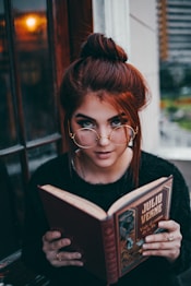A person with red hair and glasses is reading a book titled 'Julio Verne Viaje al centro de la Tierra.' The setting appears cozy, with a warm light reflecting off a window beside them. The individual is dressed in a black sweater, and there's a slight focus on their intense gaze.