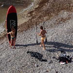 Two people stand on a pebbly beach near the shoreline, preparing for stand-up paddleboarding. One person holds a large red and black paddleboard, while the other holds two paddles. The beach is covered in small stones and some seaweed, with waves gently lapping at the shore.