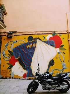 A colorful mural on a beige wall depicts a figure in a striped shirt, surrounded by symbolic elements like roses and barbed wire, accompanied by text in Spanish. In front of the mural, a black motorcycle is parked on a paved surface. The scene includes greenery from a plant on a nearby balcony.