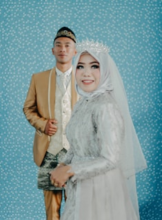 A bride in a white and silver embellished gown and a crown smiles in the foreground, with a groom in a gold suit and traditional hat standing behind her against a blue mosaic tiled background.