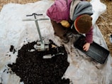 A person wearing a pink jacket and brown hat is crouching on a tarp-covered ground, preparing soil. There is a metal soil blocker tool and a pile of dark soil. The individual is placing soil into a rectangular tray using their hand. A small trowel is visible near the soil pile.
