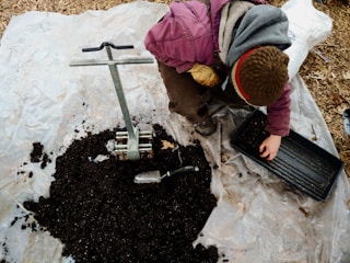 A person wearing a pink jacket and brown hat is crouching on a tarp-covered ground, preparing soil. There is a metal soil blocker tool and a pile of dark soil. The individual is placing soil into a rectangular tray using their hand. A small trowel is visible near the soil pile.