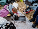 A person wearing a brown hat and purple jacket is crouched down on a plastic sheet, planting seeds in a black tray filled with soil. A wheelbarrow filled with soil stands nearby on a background of wood chips, while another person in blue jeans and black boots stands close by.