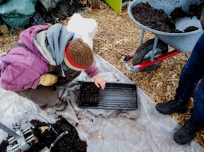 A person wearing a brown hat and purple jacket is crouched down on a plastic sheet, planting seeds in a black tray filled with soil. A wheelbarrow filled with soil stands nearby on a background of wood chips, while another person in blue jeans and black boots stands close by.
