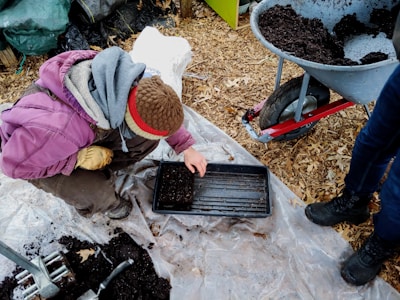 A person wearing a brown hat and purple jacket is crouched down on a plastic sheet, planting seeds in a black tray filled with soil. A wheelbarrow filled with soil stands nearby on a background of wood chips, while another person in blue jeans and black boots stands close by.