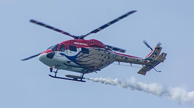 A helicopter with intricate peacock feather design on its body and a red top is flying in the sky. It belongs to the Indian Air Force, as indicated by the markings on the side. The helicopter is emitting a trail of smoke from its tail.
