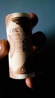 A rolled banknote held between fingers against a dark background. The note displays the denomination of 10 in an Indian currency, with intricate patterns and designs typical of currency notes.