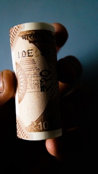 A rolled banknote held between fingers against a dark background. The note displays the denomination of 10 in an Indian currency, with intricate patterns and designs typical of currency notes.