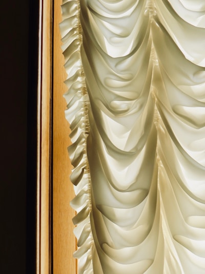 Elegant, ruched fabric drapes with a silky, luxurious texture are pulled to the side, partially revealing a wooden frame. The soft folds create a dynamic visual effect with highlights and shadows.