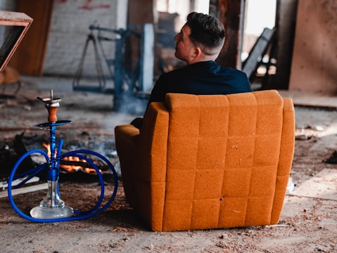 A person is sitting in an orange cushioned chair in a rustic setting with exposed beams and some debris on the ground. A blue hookah stands nearby, with its hose resting on the floor. In the background, there are signs of a small fire amid the abandoned surroundings.