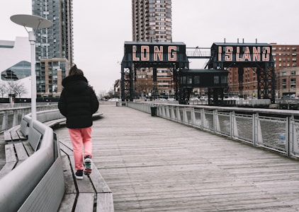 A person wearing a black jacket and pink pants walks along a wooden boardwalk with urban buildings in the background. Large industrial-style signs with the words 'LONG ISLAND' are visible, supported by metal structures. The scene appears to be overcast, with the sky covered in clouds.