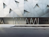 The image features a modern building facade with geometric patterns in shades of gray and white. In front of the building, large, stylized letters spell 'MIAMI'. The structure has a contemporary architectural style with angular lines and a minimalistic design. The sidewalk and street in the foreground are visible, suggesting an urban setting.