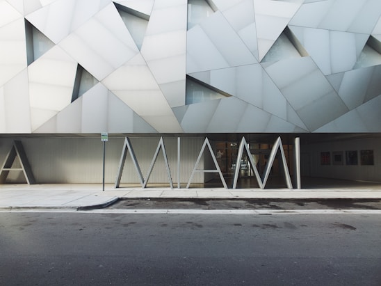 The image features a modern building facade with geometric patterns in shades of gray and white. In front of the building, large, stylized letters spell 'MIAMI'. The structure has a contemporary architectural style with angular lines and a minimalistic design. The sidewalk and street in the foreground are visible, suggesting an urban setting.