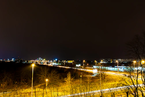 Nighttime shot of a semi-truck navigating a well-lit interstate with city lights in the background.