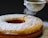 A freshly baked cake is being dusted with powdered sugar using a fine mesh sieve. The cake has a ring shape with a golden-brown crust and a soft, light texture. The powdered sugar gracefully falls onto the surface, adding a thin layer of sweetness.