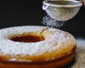 A freshly baked cake is being dusted with powdered sugar using a fine mesh sieve. The cake has a ring shape with a golden-brown crust and a soft, light texture. The powdered sugar gracefully falls onto the surface, adding a thin layer of sweetness.