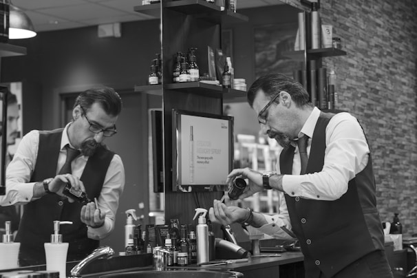 A man in a barber shop or salon is pouring some type of grooming product into his hand while looking in the mirror. The setting is a well-organized salon with various hair and grooming products neatly arranged on shelves. He is wearing a white shirt with rolled-up sleeves, a tie, and a vest, giving a professional appearance.
