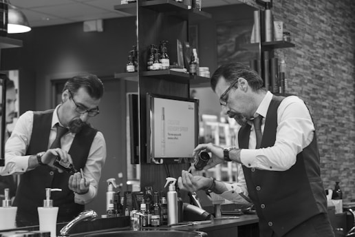A man in a barber shop or salon is pouring some type of grooming product into his hand while looking in the mirror. The setting is a well-organized salon with various hair and grooming products neatly arranged on shelves. He is wearing a white shirt with rolled-up sleeves, a tie, and a vest, giving a professional appearance.