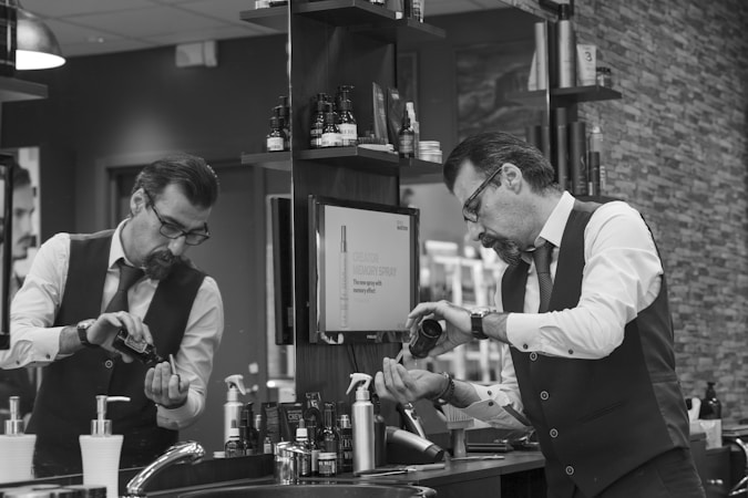 A man in a barber shop or salon is pouring some type of grooming product into his hand while looking in the mirror. The setting is a well-organized salon with various hair and grooming products neatly arranged on shelves. He is wearing a white shirt with rolled-up sleeves, a tie, and a vest, giving a professional appearance.