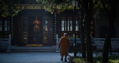 A person in traditional attire walks towards an ornate temple entrance, surrounded by trees casting shadows. The setting conveys a sense of serenity and retreat into a sacred space.