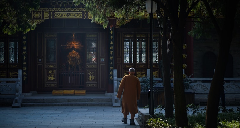 A person in traditional attire walks towards an ornate temple entrance, surrounded by trees casting shadows. The setting conveys a sense of serenity and retreat into a sacred space.