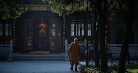 A person in traditional attire walks towards an ornate temple entrance, surrounded by trees casting shadows. The setting conveys a sense of serenity and retreat into a sacred space.
