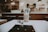 A minimalistic cafe setting with a focus on a cup of iced coffee placed on a table. The background reveals a counter, shelves with plants and items, and a blurred person. Natural lighting creates a cozy and inviting atmosphere.