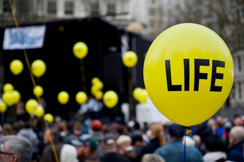 A large group of people gathered outdoors, with several yellow balloons floating above the crowd. The most prominent balloon in the foreground has the word 'LIFE' printed in bold black letters.